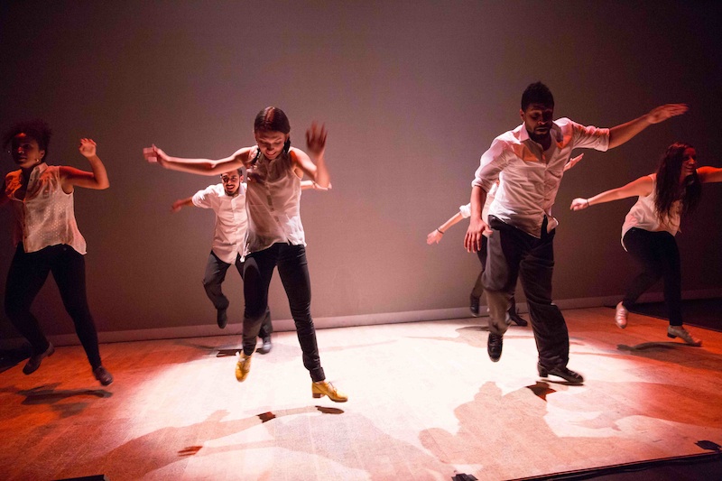 An ensemble of tappers with their arms outstretched to their sides, each are hovering over the ground